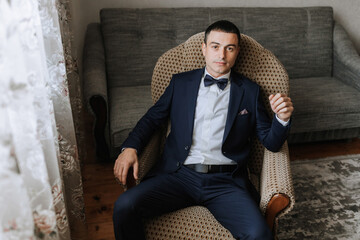 A man in a suit and bow tie sits in a chair. He is wearing a tie and a pocket square