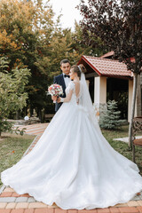 A bride and groom are standing in front of a red house with a white dress and a white veil