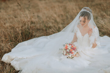 A bride is sitting in a field with a bouquet of flowers in her lap. She is wearing a veil and a...