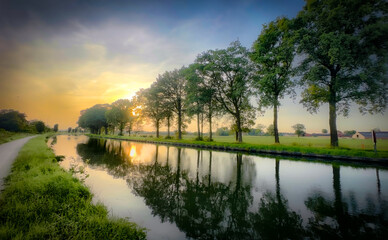 A peaceful sunset casts a warm glow over a serene canal flanked by vibrant trees, creating a tranquil scene