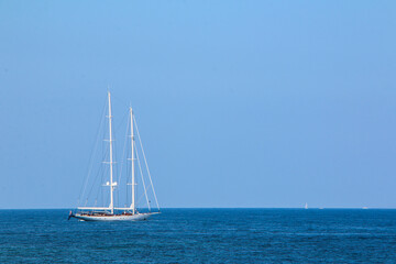 Obraz premium Sailboat with tall white masts floats on the calm Mediterranean Sea under a clear blue sky. Minimalist composition with open water and distant horizon.