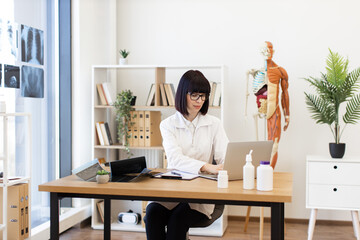 Female doctor focused on using laptop in modern medical office. Anatomical model, X-rays, and...