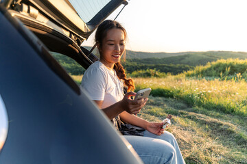 a young woman is sitting in the trunk of her car, having refreshments and checking her phone after a long hiking trip © DusanJelicic