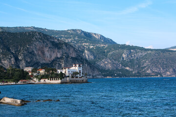 A grand villa stands on a rocky coast in Beaulieu-sur-Mer, French Riviera, surrounded by clear blue waters and mountainous terrain. Villa features classic Mediterranean architecture and lush greenery