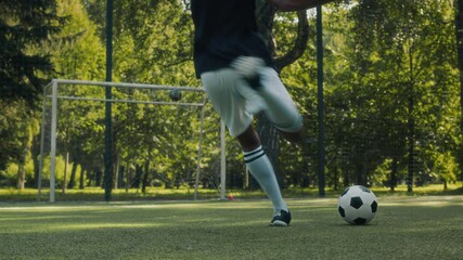 Back view shot of African American young boy wearing sports uniform kicking two soccer balls at net during outdoor practice on grassy field in summer public park