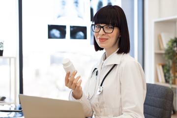 Confident female doctor holding medication bottle while sitting in modern medical office with...