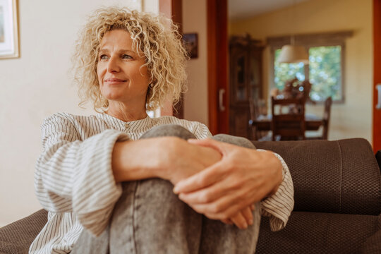 Beautiful smiling relaxed middle aged woman relaxing on sofa at home. Stylish middle aged 50s lady with curly hair, sitting on couch holding a pillow in modern living room.