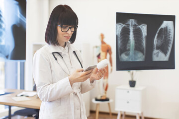 Female doctor studying x-rays of patient on glass board while writing electronic prescription on smartphone. Doctor wearing lab coat and stethoscope, working in medical office.