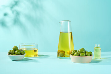Front view of glass bottles containing olive oil with bowls of fresh olives decorated on blue background. Natural cosmetic research concept with olive extract