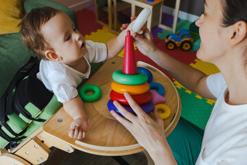 Handsome little boy with muscle weakness is actively participating in a rehabilitation session, focusing on tactile play with colorful stacking rings
