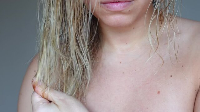 Young caucasian blonde woman applying oil with vitamins on her split ends using a dropper rubbing it to dry hair on a dark grey background. Hair care
