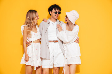 Group of young three stylish friends posing in studio. Fashion man and two cute female dressed in casual summer clothes. Smiling models having fun. Cheerful women and guy near yellow wall, sunglasses