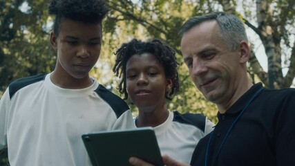 Tilt up of Caucasian adult male soccer coach showing video of match to young African American teammates while standing on outdoor field during training session - Powered by Adobe