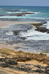 A scenic view of unique rock formations along a sandy beach at low tide, with gentle waves breaking in the background. The image captures the natural beauty and tranquility of the coastal landscape