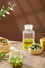 Minimal scene - fresh olives on wooden bowls, olive oil and green leaves decorated on brown background. Minimal concept for advertising product with ingredient from olive