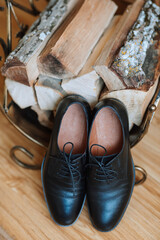 Two black shoes are on a wooden table next to a pile of wood. The shoes are tied together with a shoelace