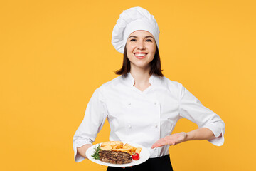 Young smiling chef cook baker woman wear white shirt black apron uniform toque chefs hat hold in hand point on plate ready dish steak isolated on plain yellow background studio. Cooking food concept.