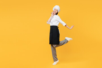 Full body side view smiling young chef cook baker woman wear white shirt black apron uniform toque hat raise up leg put hand on face isolated on plain yellow background studio. Cooking food concept.