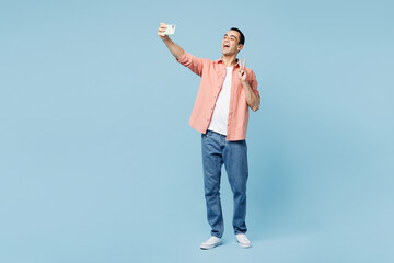 Full body young middle eastern man he wear pink shirt white t-shirt doing selfie shot on mobile cell phone post photo on social network isolated on plain light blue cyan background. Lifestyle concept.
