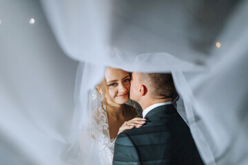 A bride and groom are hugging each other under a white veil. The bride is wearing a white dress and the groom is wearing a suit. Scene is happy and romantic