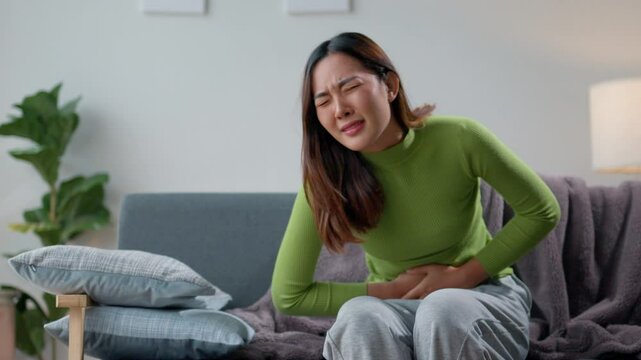Woman experiencing painful cramps while lying on a sofa in her living room. She is holding her belly and grimacing in pain