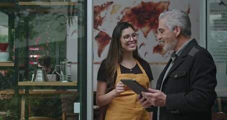 Man discussing with female barista wearing a yellow apron outside a coffee shop. They are both looking at a tablet screen, exchanging ideas and information. Coffee brewing equipment in the foreground - Powered by Adobe