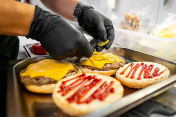 A close-up of hands in black gloves preparing cheeseburgers with condiments on a grill. The focus is on the food preparation process, highlighting the ingredients and the act of cooking.