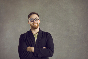 Studio shot of happy nerd. Portrait of funny guy isolated on grey text copy space background. Bearded young man in suit jacket and round thick lens glasses standing arms folded and looking at camera