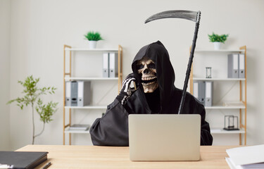 Devil employee in black cloak and skeleton mask sitting at workplace in office. Skeleton in grim...