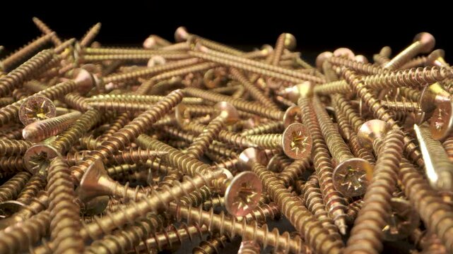 Close up view of a pile of gold screws. The screws are arranged in a random pattern and are illuminated by a bright light. The video captures details of the screws, including the threads and the heads