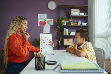 Teacher with long braided hair pointing at colorful alphabet chart while young student in striped...