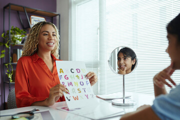Fototapeta premium Female therapist engaging in speech therapy session with young child holding alphabet card. Child's enthusiastic reflection seen in circular mirror, room filled with educational tools