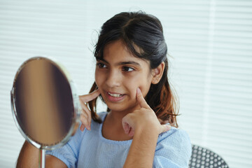 Young girl with dark hair smiling at her reflection while gently touching her cheeks in a bright room with white blinds and simple furniture