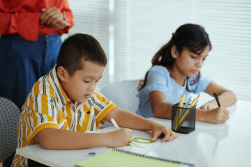 Young students concentrating on completing classroom writing assignments in organized setting with teacher supervising. Natural lighting brightens the room, fostering a conducive learning environment