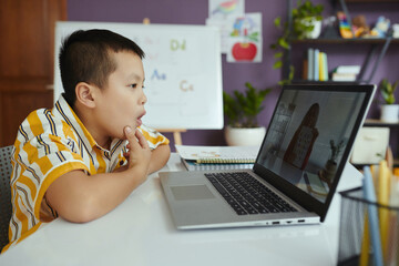 Young Asian boy attentively engaging in a virtual class, with a laptop placed on a table and bookshelves in the background. Online learning environment with educational elements in view