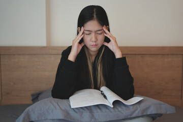 Young asian teenager struggle doing homework with tablet and notebook while sitting on bed, learning at home, and getting headache. People struggle with technology and try hard to pass the exam.