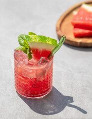 Glass of refreshing watermelon cocktail with basil and ice with fruit slices and fresh herb on grey concrete background with shadow.