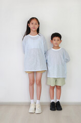 Asian young girl child and little boy kid standing and looking at camera against white wall in the room. Sister and brother relationship. Full body photo.