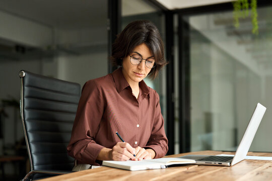 Busy young Hispanic business woman company owner ceo working in office using laptop writing notes. Serious female professional executive manager in her 30s using computer, elearning at workplace.