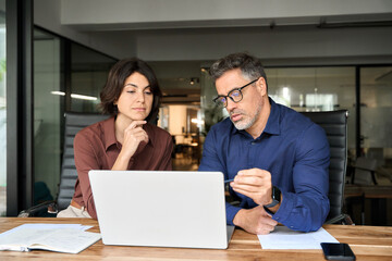 Two busy professional executives team using laptop in corporate office. Business man and woman leaders managers working on computer at work desk having conversation on financial project at meeting.