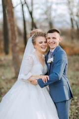 Fototapeta premium A bride and groom are posing for a picture in a forest. The bride is wearing a white dress and the groom is wearing a blue suit. They are both smiling and seem happy