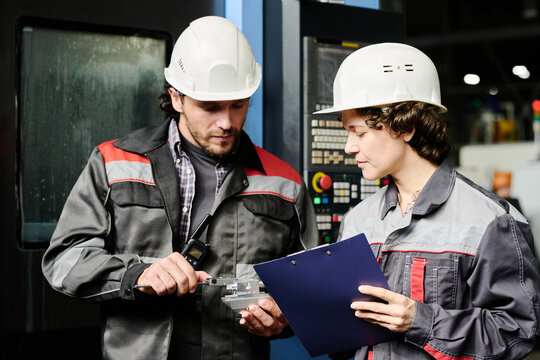 Two engineers in hard hats discussing machinery setup in industrial facility while holding technical documents and device