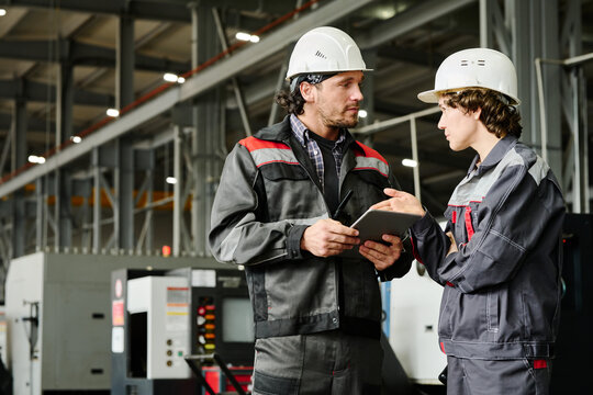Two engineers collaborating in industrial workshop, examining tablet and discussing project surrounded by machinery and workspace equipment