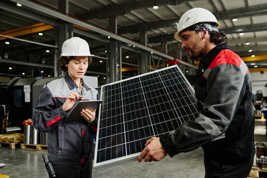 Two workers in a factory setting inspecting and documenting solar panels while wearing protective gear and helmets