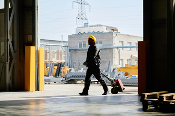 Worker moving loaded hand truck in industrial warehouse during daytime with visible heavy machinery and industrial equipment in the background