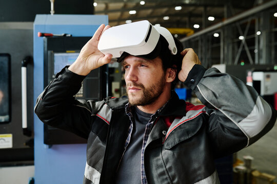 Man in industrial setting wearing virtual reality headset adjusting device, surrounded by machinery in spacious facility with metal structures