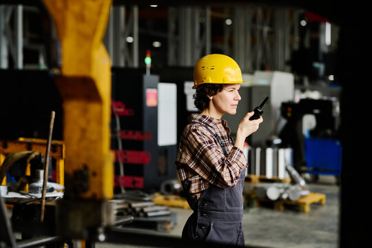 Young engineer wearing hard hat, holding radio device while standing in industrial workshop