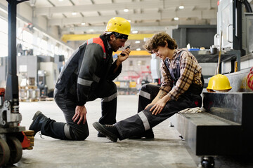 Two workers wearing protective gear are discussing mechanical work in an industrial workshop setting filled with equipment and machinery
