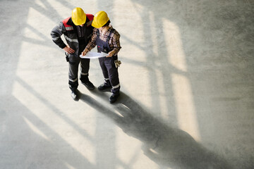 Construction workers standing on site reviewing blueprints and discussing project details in sunlight shadows