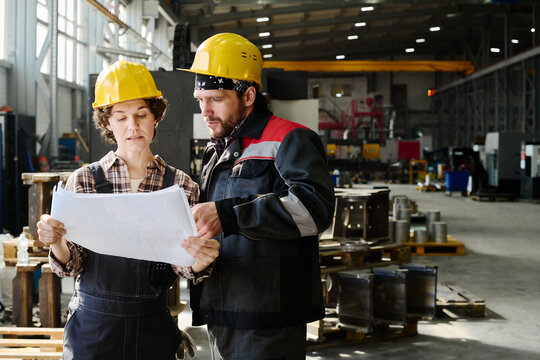 Two factory workers in hard hats examining paper blueprints indoors in manufacturing facility surrounded by machinery and metal parts